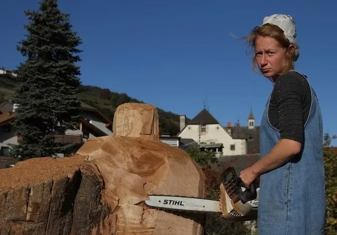 Clara Mayr carving a large block of wood with a chainsaw, outdoors in a mountain village
